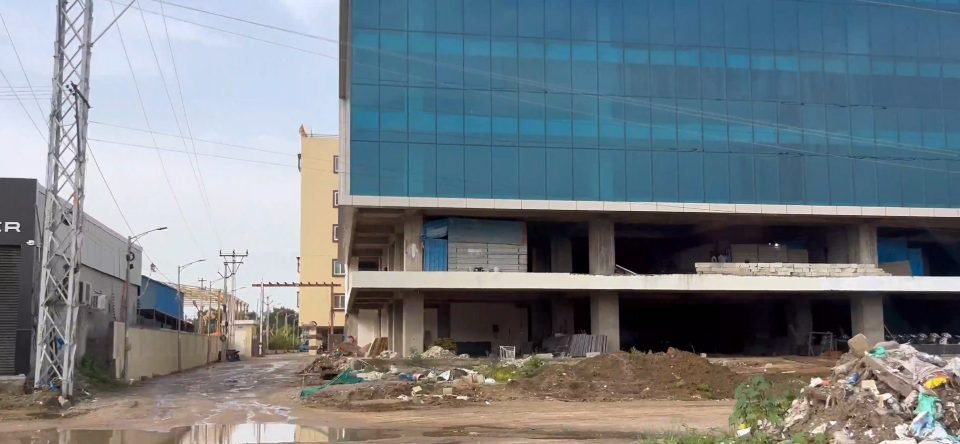A multi-storey glass-fronted building under construction with an exposed lower floor, beside a wet service road and a power tower