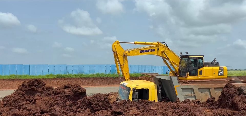 A Komatsu PC300 LC excavator loading a yellow tipper truck with red soil, in front of a long blue sheet-metal boundary fence