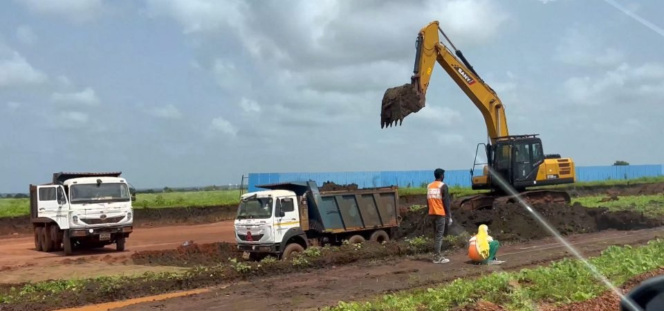 A Sany excavator lifting soil beside two white tipper trucks, with workers in safety vests and a blue boundary fence behind