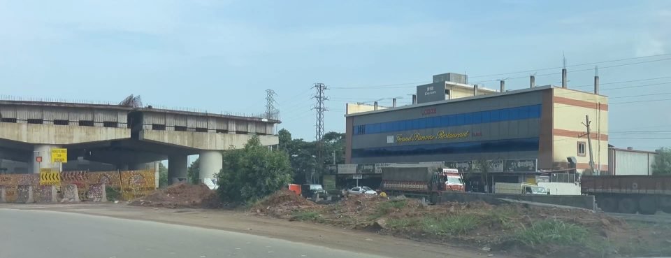 An unfinished flyover deck with a road-closed diversion sign, beside a three-storey roadside restaurant building and parked lorries