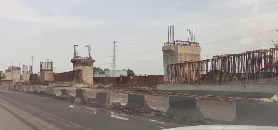 Concrete piers of an elevated highway structure with exposed reinforcement bars, behind concrete barriers beside an active road