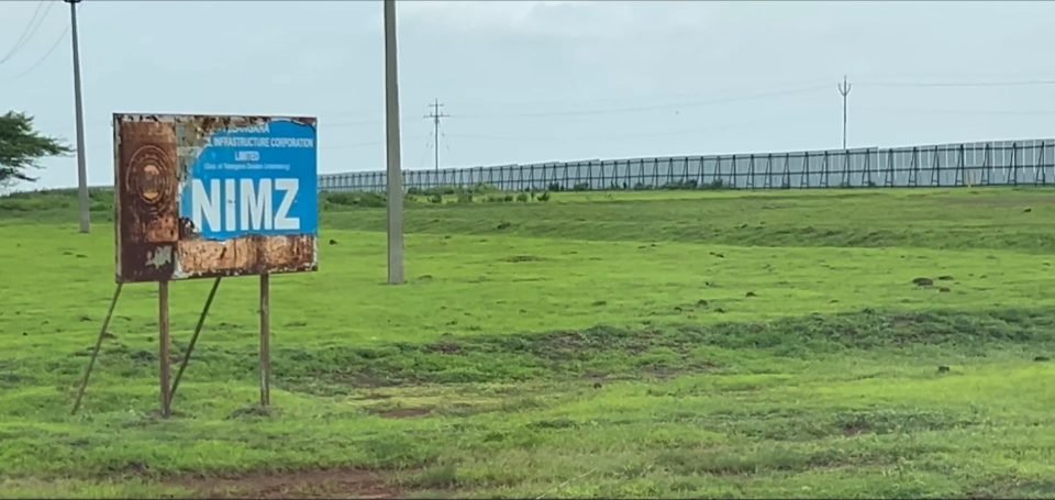 A rusted, peeling NIMZ signboard of the Telangana State Industrial Infrastructure Corporation standing in green grassland, with a long fence line behind