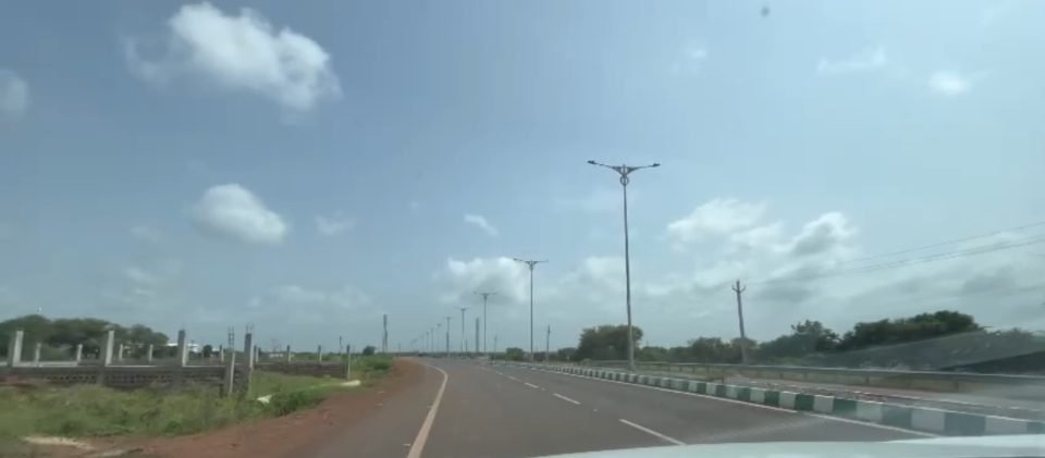 A newly built multi-lane road at the NIMZ perimeter with a median, tall street-light poles, a fence line and no traffic or people