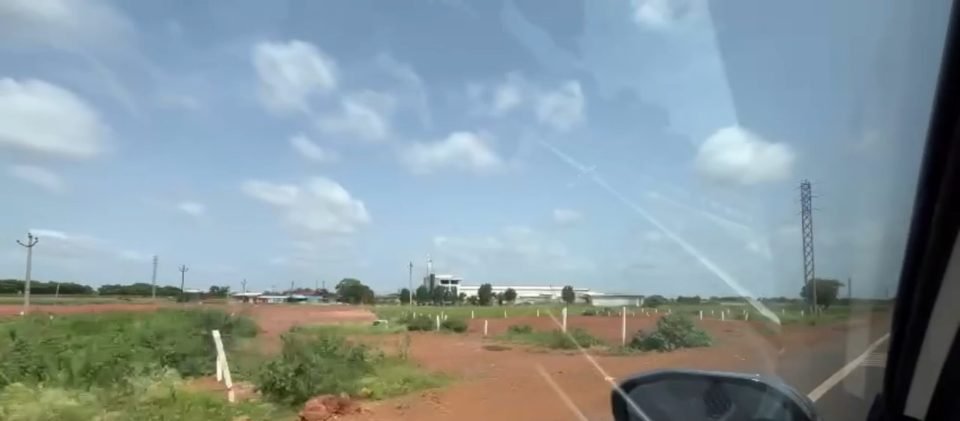 Cleared red-soil land with boundary posts and scrub, and low industrial buildings on the horizon, seen from a car window
